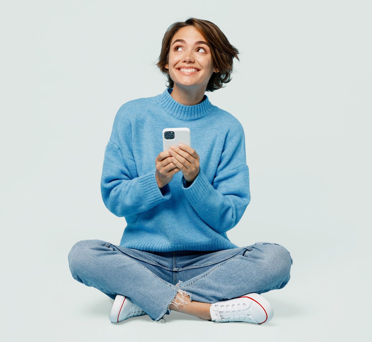 A smiling woman sitting cross-legged in a blue jumper, holding her phone